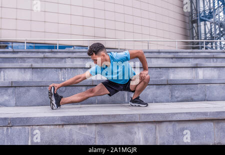 Male athlete, tanned man, summer in city, stretching muscles of legs, knees, ankles, warming up before jogging, fitness training, workout, concrete Stock Photo