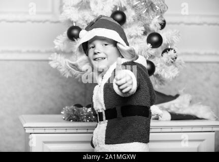 Christmas tree ideas for kids. Boy kid dressed as cute elf magical creature white artificial ears and red hat near christmas tree. Christmas elf costume for child. Christmas party with elf costume. Stock Photo