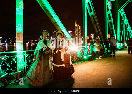 27 September 2019, Hessen, Frankfurt/Main: Two women in costumes from the Biedermeier period stand on the colourfully illuminated Iron Bridge. The pedestrian bridge was opened 150 years ago and the steel structure has become one of the city's landmarks. Photo: Frank Rumpenhorst/dpa Stock Photo