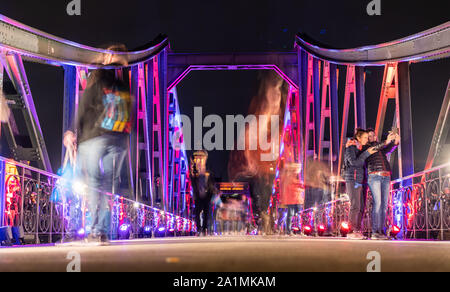 27 September 2019, Hessen, Frankfurt/Main: Passers-by cross the Main River on the colourfully illuminated Iron Bridge. The pedestrian bridge was opened 150 years ago and the steel structure has become one of the city's landmarks. Photo: Frank Rumpenhorst/dpa Stock Photo