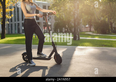 Brunette girl riding an eco-friendly electric kick scooter in a park in sunny weather on sidewalks Stock Photo