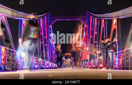 27 September 2019, Hessen, Frankfurt/Main: Passers-by cross the Main River on the colourfully illuminated Iron Bridge. The pedestrian bridge was opened 150 years ago and the steel structure has become one of the city's landmarks. Photo: Frank Rumpenhorst/dpa Stock Photo