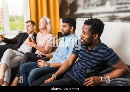 Young handsome bearded african american guy watching interesting exciting movie on tv at home with his multiethnic friends, drinking soft beverages. S Stock Photo