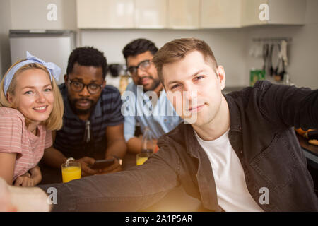 Say Cheese. Handsome caucasian man taking selfie with friends. Happy multi ethnic students sitting at table and having lunch in kitchen at home. Stock Photo