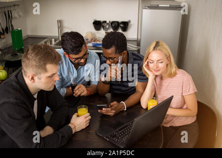 Top view of four multiracial students sitting at table, drinking soft beverages, using smartphones, watching video on laptop together. Group of friend Stock Photo