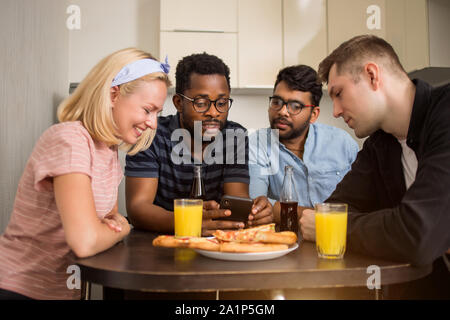 Four friends sitting at table, eating pizza, drinking soft beverages, reading text message on smartphone, testing new phone application together. Mult Stock Photo