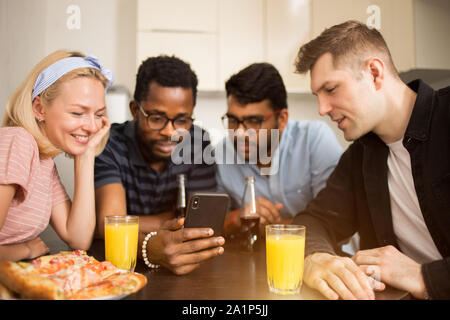 Three young man and woman sitting in kitchen, using mobile phone together while eating pizza. Friends playing games on smartphone, watching funny vide Stock Photo
