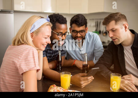 Beautiful happy young caucasian blonde woman sitting at table with her friends, laughing looking at smartphone. Multi ethnic students testing new gadg Stock Photo