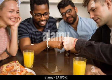 Group of four students having fun in kitchen. Young african and caucasian men giving fist bump, smiling celebrating success with friends. Friendship, Stock Photo