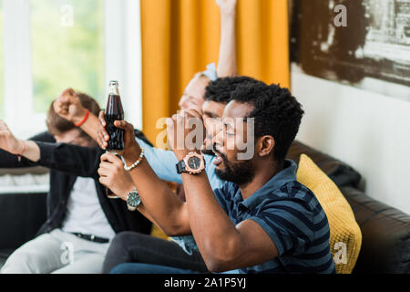 Side view shot of young african bearded man watching sport match with friends, cheering up for favorite team, clenching fists, sitting with drinks on Stock Photo