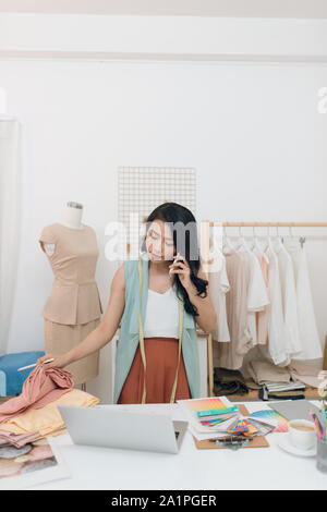 Discussing fashion trends. Beautiful young woman talking on the mobile phone and working on laptop while standing in her workshop Stock Photo