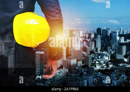 business man in suit or project manager holding yellow safety helmet with during sunrise and cityscape background, real estate business Stock Photo