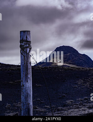 An old withered wooden pole with rusty barbed wire in front of a dark grey sky and a large rocky mountain Stock Photo