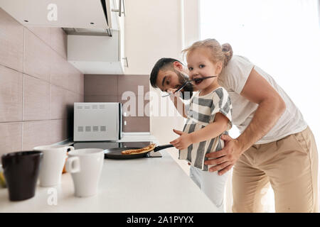 hardworking little girl helping her father to prepare pancakes, looking at the camera while standing nex to cooker. close up side view photo. copy spa Stock Photo