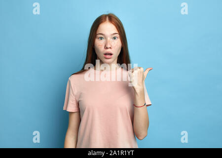 beautiful emotional freckled girl with ginger hair, has surprised expression, indicates aside, shows free space for advertisement.isolated blue backgr Stock Photo