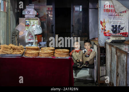 Xian, China -  August 2019 :  Two little boys playing games on a small mobile device while sitting on chairs in a small local bakery on the street in Stock Photo