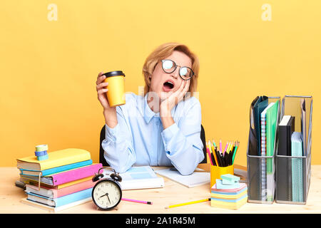 unhappy sad depressed woman with wide open mouth yawning while sitting at workplace. boring work, close up portrait, isolated yellow background, studi Stock Photo