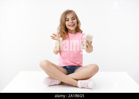 Cheerful tender blond lively enthusiastic young teenage girl, sit crossed legs, hold smartphone, record video-blog become blogger, gesturing excited Stock Photo