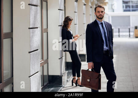 handsome cute businessman wearing a suit and holding a brown briefcase . financial concept.young attractive woman using tablet in the blurred backgrou Stock Photo
