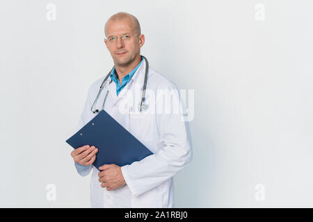 Young successful bald practitioner in whitecoat holding document while standing in front of camera by wall Stock Photo