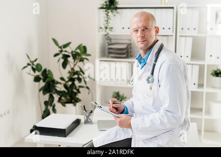 Handsome doctor in whitecoat sitting on desk and looking at you while making medical notes or prescriptions Stock Photo