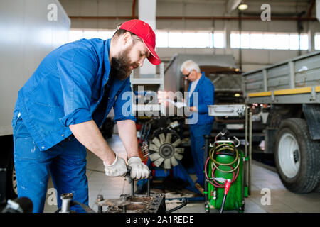 Bearded man in workwear fixing one of engine details while repairing on background of his master or colleague Stock Photo