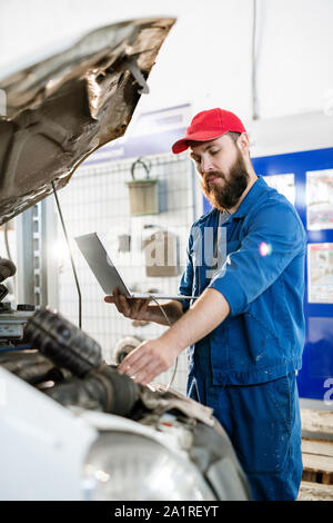 Young bearded technician of repair service examining broken engine of lorry and using laptop to find online information Stock Photo