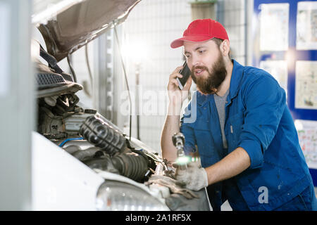 Bearded young worker of machine repair service talking to client on mobile phone while standing by engine of broken car Stock Photo