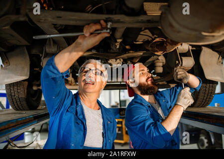 Young and mature technicians of modern car repair service fixing details of car motor while standing under machine Stock Photo