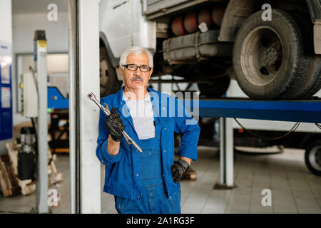 Serious grey-haired man in workwear standing in workshop or hangar in front of camera before starting to work Stock Photo