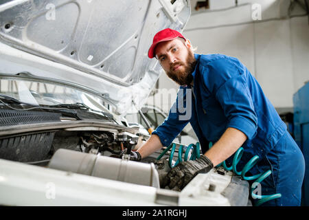 Serious technician in workwear looking at you while repairing engine of broken machine in his workshop or hangar Stock Photo