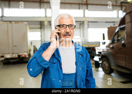Senior professional technician in workwear and eyeglasses standing in workshop and consulting client on mobile phone Stock Photo