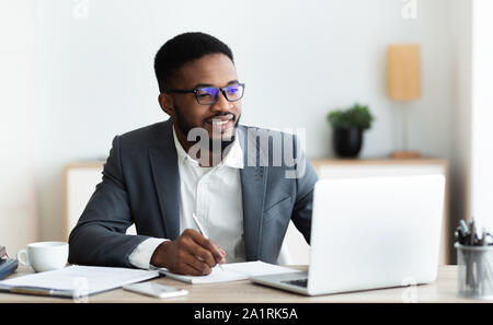 Handsome black entrepreneur working on laptop and taking notes Stock Photo