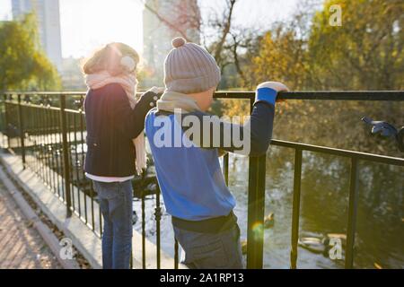 Children boy and girl standing backs on bridge, looking at ducks, sunny autumn day in park, golden hour Stock Photo