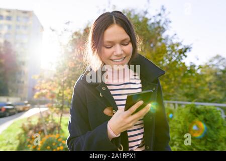 Teen girl in coat using smartphone, sunny autumn day, golden hour Stock Photo