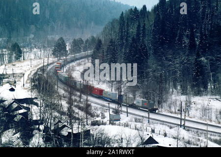 freight train moving through coniferous forest during snowfall along frozen river among hills. Northern country. Side fron top Stock Photo