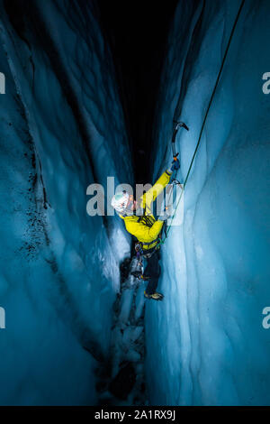 Athlete ice climbing inside a crevasse at night, lit by artificial light as he climbs from the dark crevasse. Stock Photo