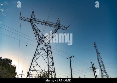 View of an electric power tower in the foreground. Beside, a data communication tower and a power pole. The setting sun hidden behind illuminates the Stock Photo