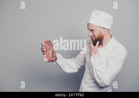 Profile side view portrait of nice handsome minded butcher guy holding fresh meat in arm hand thinking how to cook prepare touching chin beard Stock Photo