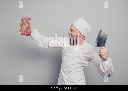 Portrait of he crazy mad dangerous attractive handsome butcher guy holding fresh meat and knife in arms hands opened mouth isolated over gray pastel Stock Photo