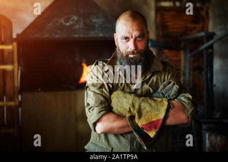 Portrait of a brutal, confident blacksmith, a man with a beard. Standing in the workshop, folding his arms over his chest. Stock Photo