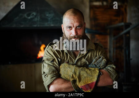 Portrait of a brutal, confident blacksmith, a man with a beard. Standing in the workshop, folding his arms over his chest. Stock Photo