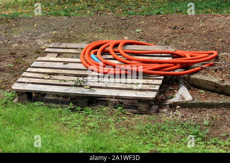 Broken wooden pallet with orange plastic hose on top surrounded with grass and fallen leaves in local urban garden on warm sunny summer day Stock Photo