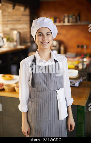 Portrait of cheerful young cook wearing chefs hat and apron smiling at camera while standing in restaurant kitchen Stock Photo