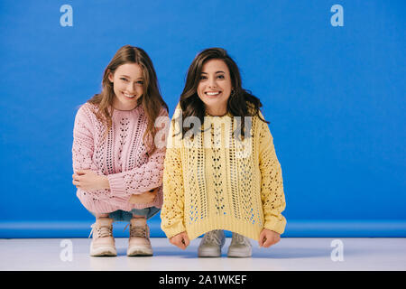 attractive and smiling women in sweaters looking at camera on blue background Stock Photo