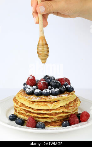 Woman pouring honey over a stack of pancakes with blueberries and red raspberries dusted with icing sugar on a white background Stock Photo