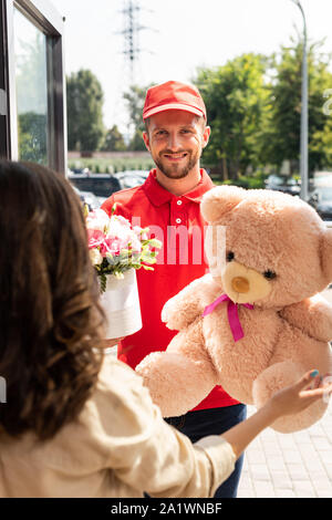 selective focus of cheerful delivery man in cap holding teddy bear and flowers near woman Stock Photo