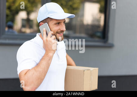 happy bearded delivery man talking on smartphone while holding box Stock Photo