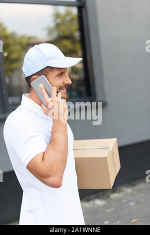 cheerful and bearded delivery man talking on smartphone while holding box Stock Photo