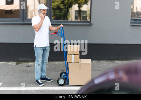 selective focus of happy bearded delivery man talking on smartphone while holding delivery cart Stock Photo